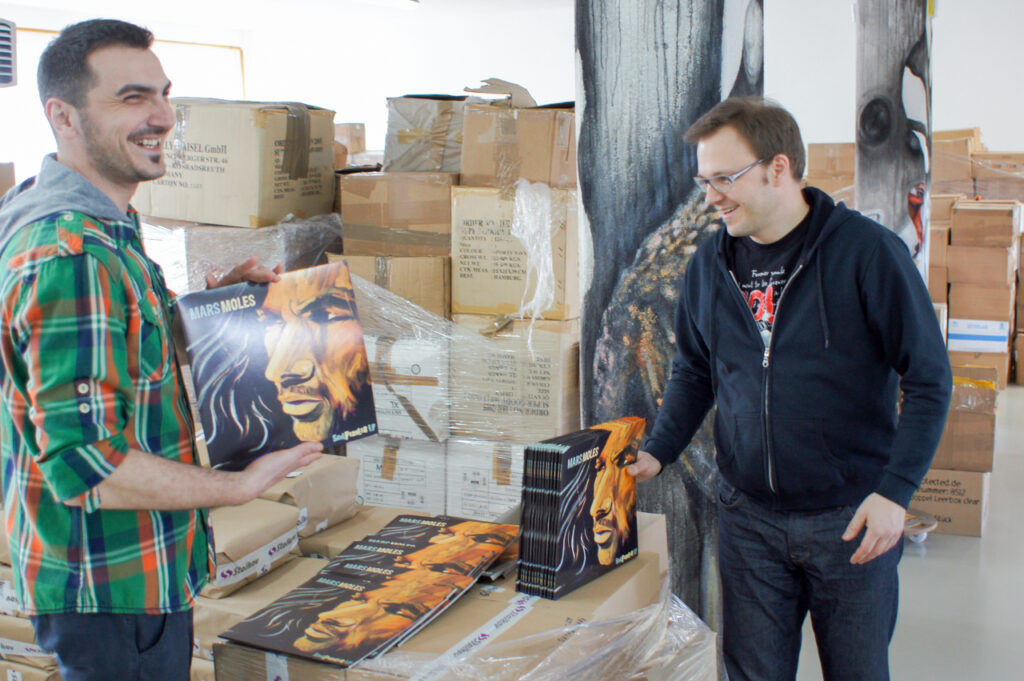 Zwei Männer prüfen sorgfältig Vinyl-Schallplatten des Künstlers \"Mars Moles\" in einer Lagerhalle, vermutlich während der Vinylherstellung.